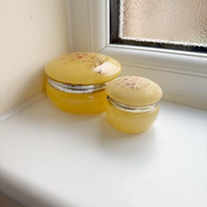 May include: Two yellow, oval-shaped trinket boxes with silver rims and lids. The lids are decorated with a floral design in shades of red and brown. The boxes are sitting on a white windowsill.