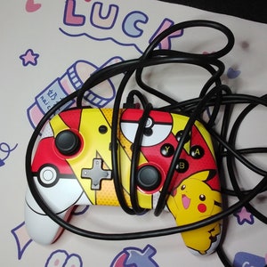 May include: A black and yellow video game controller with a Pikachu design. The controller has a red, yellow, and black Poké Ball design on the back. The controller is on a pink and white background with a black cable.