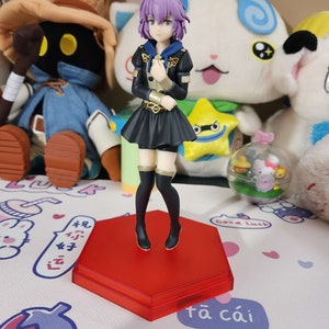 May include: A purple-haired anime figurine with a black dress and gold accents standing on a red hexagonal base. The figurine is posed with hands clasped in front of her.