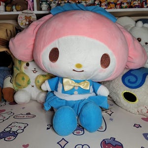 May include: A pink and white plush toy with a blue dress and yellow bow. The toy has large, brown eyes and a smiling mouth. The toy is sitting on a white surface with other plush toys.