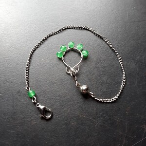 May include: A silver-colored nipple ring with a chain and clasp. The ring features a cluster of five small, round, green beads. A single green bead is attached to the chain near the clasp. The jewelry is displayed against a dark background.