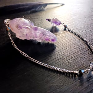 May include: A handcrafted amethyst pendant with a silver chain. The pendant is a carved crystal with a chain and a small amethyst bead. A black feather rests in the background. The jewelry is on a dark surface.