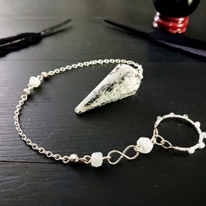 May include: A silver-toned pendulum with a chain, a cone-shaped crystal, and a ring. The crystal is white with black markings. The ring is adorned with small, clear beads. The pendulum is on a dark surface.