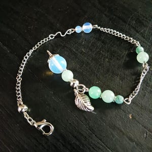 May include: A delicate silver bracelet featuring a chain design, adorned with light blue and green beads. A silver leaf charm and a larger blue bead pendant add detail. The bracelet has a clasp closure.