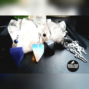 May include: A collection of crystal pendulums with silver chains. The pendulums are various colors, including blue, white, and black. The background features a large white crystal and the text "SPIRAL CROW PENDULUMS" in a circular logo.