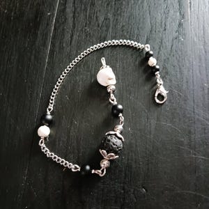 May include: A silver chain bracelet featuring a white skull charm, black and white beads, and a lava rock bead. The bracelet has a lobster clasp closure. The beads are strung on a silver chain.