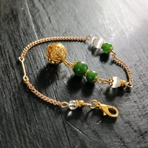 May include: A gold-colored bracelet featuring a chain and decorative elements. The bracelet includes a filigree sphere, green beads, and clear and white beads. It has a lobster clasp closure.