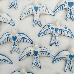Puede incluir: Una colección de adornos de pájaros de cerámica blanca con detalles pintados en azul. Cada pájaro tiene un corazón azul en el pecho y patrones intrincados en sus alas.