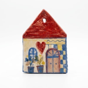 Könnte beinhalten: Ein flaches, hausförmiges Keramikornament mit rotem Dach und einem Loch zum Aufhängen. Die cremefarbene Fassade zeigt ein gemaltes rotes Herz, ein blaues Bogenfenster, zwei Topfpflanzen und eine Holztür mit blau-weißen Karomustern als Akzent.