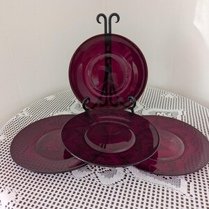 May include: A set of four burgundy-colored glass plates. One plate is displayed on a black metal plate stand. The plates have a decorative pattern and are arranged on a white lace tablecloth. The plates are a deep red color.