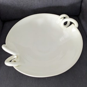 May include: A white, round serving bowl with two loop handles. The bowl has a smooth surface and a shallow depth. The handles are integrated into the design, adding a decorative element. The bowl is likely made of ceramic or porcelain.