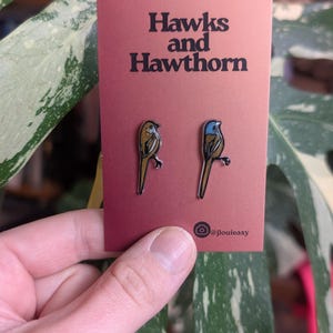 Könnte beinhalten: Zwei vogelformige Emaille-Anstecknadeln auf einer weinroten Karte mit dem Text "Hawks and Hawthorn". Eine Nadel ist goldfarben mit schwarzer Umrandung, die andere blau und goldfarben. Der Hintergrund zeigt ein grün-weißes Blatt.