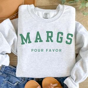 May include: A light gray sweatshirt with the text "MARGS POUR FAVOR" in green block letters. The sweatshirt is folded on top of blue denim shorts and a pair of brown sandals. A brown hat is visible in the background.