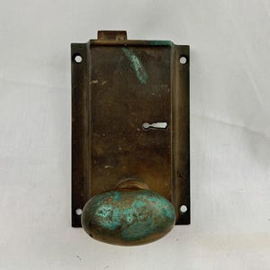 Op de afbeelding: Antiek messing deurknop met een groene patina en een sleutelgat. De knop is ovaalvormig en heeft een platte achterplaat.