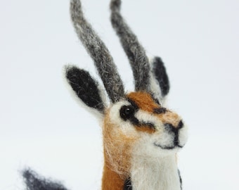May include: A handcrafted felt sculpture of a gazelle head. The gazelle features a white body, a brown and orange face, black and gray horns, and a black and white tail. The sculpture is set against a white background.
