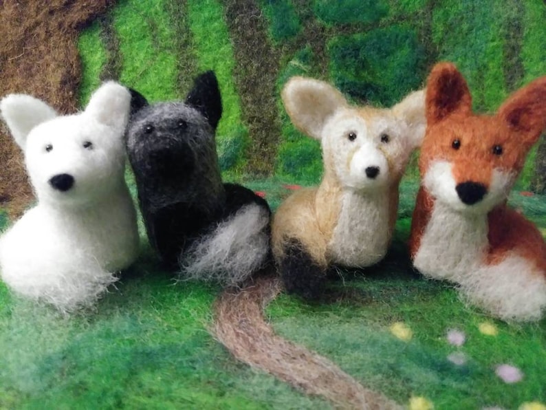 Puede incluir: Cuatro figuritas de animales de fieltro, un perro blanco, un perro blanco y negro, un zorro marr&oacute;n y un zorro naranja. Las figuritas est&aacute;n sentadas sobre una superficie verde cubierta de hierba.