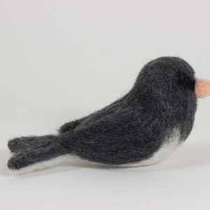 May include: A handmade felt bird figurine in shades of gray, white, and pink. The bird has a dark gray body, a white belly, and a small pink beak. The felted bird is a decorative item.