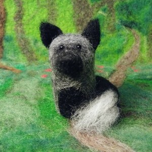 Puede incluir: Una figurita de perro de fieltro negro y blanco con una cola larga y tupida. El perro est&aacute; sentado sobre una superficie verde cubierta de hierba con un fondo borroso de &aacute;rboles.