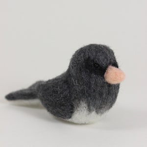 May include: A handmade felt bird figurine. The bird is primarily gray with a white chest and a small, light pink beak. The bird is in a resting position, with its tail feathers slightly extended.