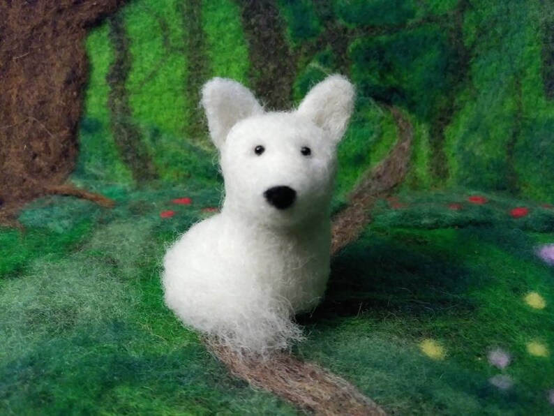 Puede incluir: Figurita de perro de fieltro de aguja blanca con ojos y nariz negros. La figurita est&aacute; sentada sobre un fondo verde y marr&oacute;n.
