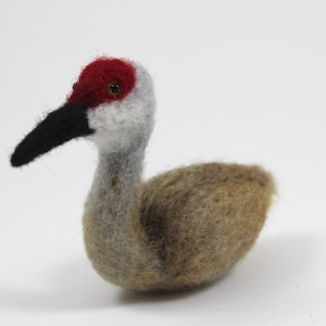 Op de afbeelding: Een handgevoelde zandhillkraanvogelfiguur met een rode kroon, een zwarte snavel en grijze en bruine veren.