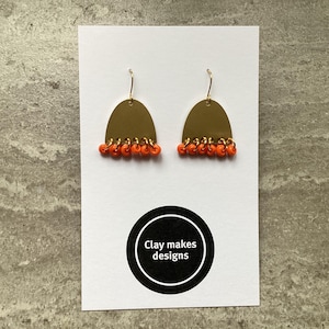 May include: Gold-coloured, half-moon earrings with orange beads suspended from the base. The earrings are presented on a white card with a black and white circular logo that reads "Clay makes designs".