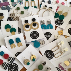 May include: Assortment of handmade polymer clay earrings on white cards with the text "Clay makes designs". The earrings feature various colours, patterns, and shapes, including geometric, abstract, and animal print designs.