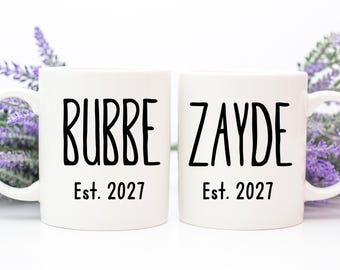 May include: Two white ceramic mugs featuring black text. The left mug displays the word BUBBE above the text Est. 2027. The right mug displays the word ZAYDE above the text Est. 2027. Both mugs have a smooth finish and a standard handle on the side.