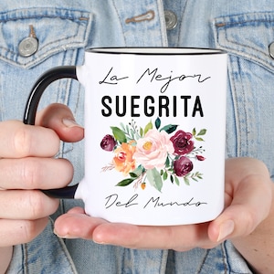 May include: White ceramic mug with a black handle and rim, featuring the text "La Mejor Suegrita Del Mundo" in black script. A floral arrangement in shades of pink, orange, and burgundy decorates the mug.