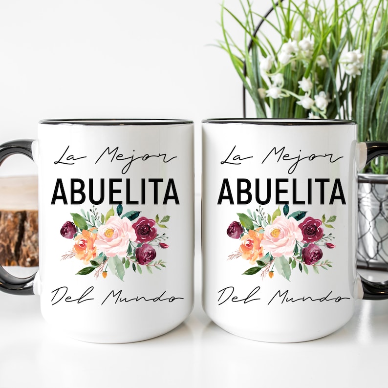 May include: Two white ceramic mugs with black handles and black rims. Both mugs feature a floral design with pink, orange, and burgundy flowers. The text "La Mejor Abuelita Del Mundo" is printed in black on each mug.
