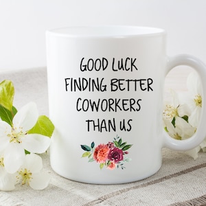 Peut inclure: Une tasse &agrave; caf&eacute; en c&eacute;ramique blanche avec du texte noir qui dit "Bonne chance pour trouver de meilleurs coll&egrave;gues que nous". La tasse a un motif floral avec des fleurs roses et rouges.