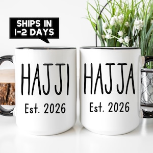May include: Two white ceramic mugs with black rims and handles. Each mug has the name "HAJJI" and "HAJJA" printed in black, along with "Est. 2026". A black banner above reads "SHIPS IN 1-2 DAYS".