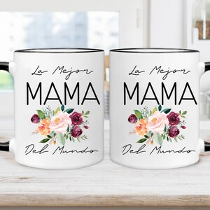 Puede incluir: Dos tazas de cer&aacute;mica blanca con bordes y asas negros. Cada taza presenta un dise&ntilde;o floral con flores rosas, naranjas y burdeos. El texto "La Mejor Mama Del Mundo" est&aacute; impreso en negro en cada taza.