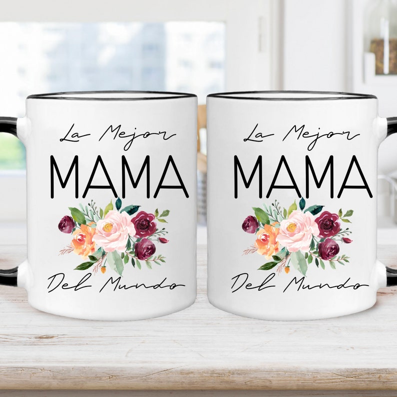 Puede incluir: Dos tazas de cer&aacute;mica blanca con bordes y asas negros. Cada taza presenta un dise&ntilde;o floral con flores rosas, naranjas y burdeos. El texto "La Mejor Mama Del Mundo" est&aacute; impreso en negro en cada taza.