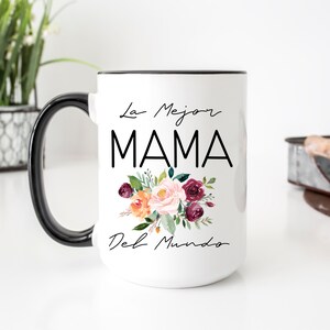 Puede incluir: Taza de cer&aacute;mica blanca con asa y borde negros. La taza presenta un dise&ntilde;o floral con flores rosas, naranjas y burdeos. El texto "La Mejor MAMA Del Mundo" est&aacute; impreso en negro en la taza.