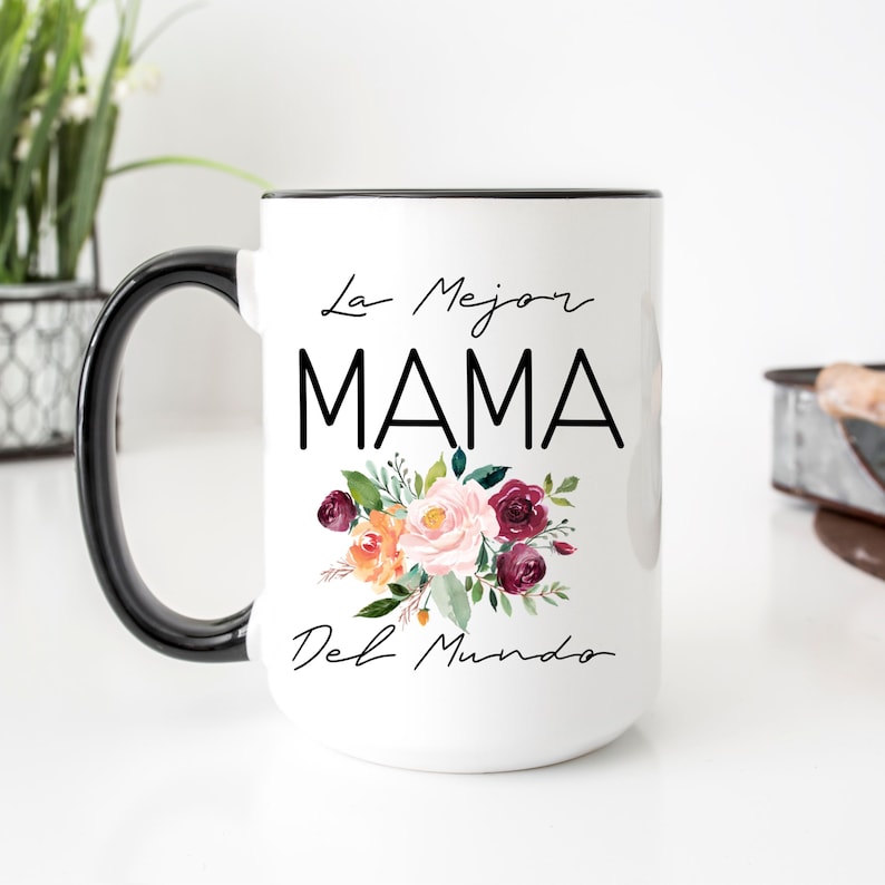 Puede incluir: Taza de cer&aacute;mica blanca con asa y borde negros. La taza presenta un dise&ntilde;o floral con flores rosas, naranjas y burdeos. El texto "La Mejor MAMA Del Mundo" est&aacute; impreso en negro en la taza.