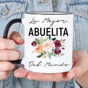 May include: White ceramic mug with a black handle and rim. The mug has a floral design with pink, orange, and burgundy flowers. The text "La Mejor Abuelita Del Mundo" is printed on the mug.