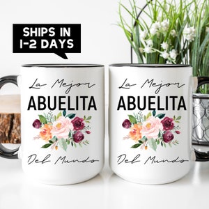 May include: Two white ceramic mugs with black rims and handles. Both mugs feature a floral design with pink, orange, and burgundy flowers. The text "La Mejor Abuelita Del Mundo" is printed in black on each mug.