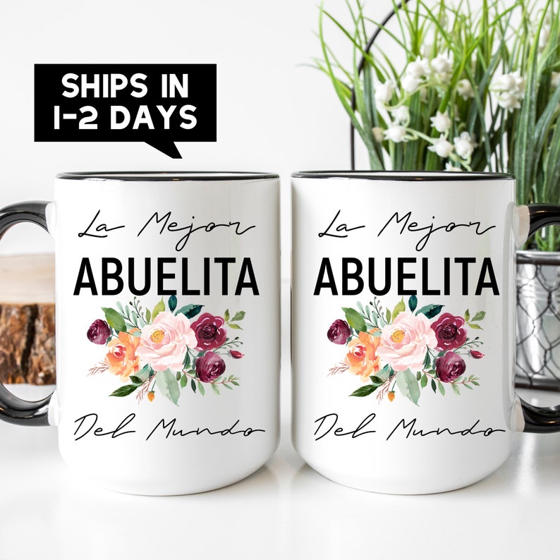 May include: Two white ceramic mugs with black rims and handles. Both mugs feature a floral design with pink, orange, and burgundy flowers. The text "La Mejor Abuelita Del Mundo" is printed in black on each mug.