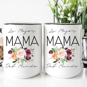 Puede incluir: Dos tazas de cer&aacute;mica blanca con bordes y asas negras. Ambas tazas presentan un dise&ntilde;o floral con flores rosas, naranjas y burdeos. El texto "La Mejor MAMA Del Mundo" est&aacute; impreso en negro en cada taza.
