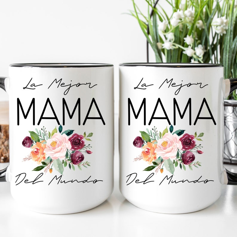 Puede incluir: Dos tazas de cer&aacute;mica blanca con bordes y asas negras. Ambas tazas presentan un dise&ntilde;o floral con flores rosas, naranjas y burdeos. El texto "La Mejor MAMA Del Mundo" est&aacute; impreso en negro en cada taza.