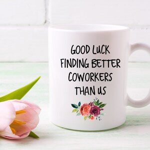 Peut inclure: Une tasse &agrave; caf&eacute; en c&eacute;ramique blanche avec du texte noir qui dit "Bonne chance pour trouver de meilleurs coll&egrave;gues que nous". La tasse a un motif floral avec des fleurs rouges, roses et orange.