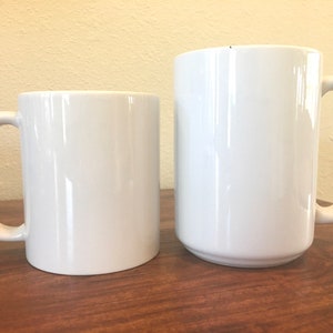 May include: Two white ceramic mugs of different sizes. The smaller mug is cylindrical, while the larger one has a slightly tapered shape. Both mugs have a curved handle and a glossy finish. They are placed on a wooden surface.