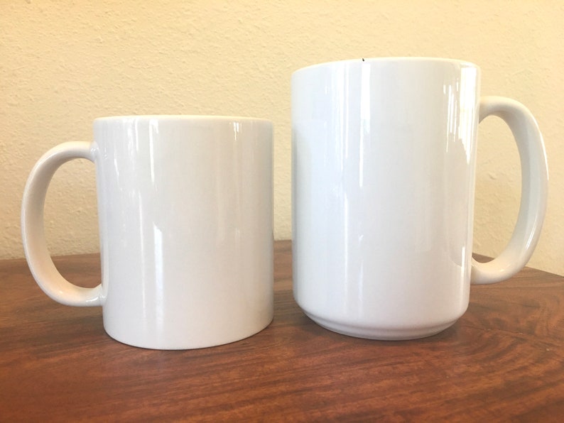 May include: Two white ceramic mugs of different sizes. The smaller mug is cylindrical, while the larger one has a slightly tapered shape. Both mugs have a curved handle and a glossy finish. They are placed on a wooden surface.