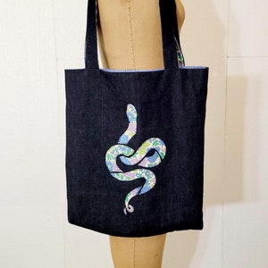Op de afbeelding: Een donkerblauwe denim tote bag met een bloemenpatroon slangenontwerp. De tas heeft een blauw-wit gestreepte voering.
