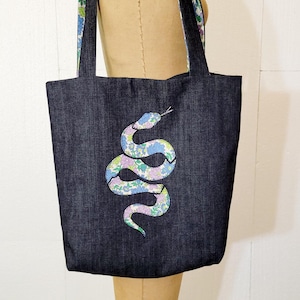 Op de afbeelding: Een donkerblauwe denim tote bag met bloemige gepatenteerde hengsels. De tas heeft een bloemig slangontwerp aan de voorkant.