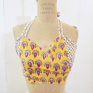 May include: A yellow halter top with a red and green floral print. The top is made of two different fabrics, one with a larger floral print and the other with a smaller geometric print. The top is tied at the neck and has a wrap-around style.