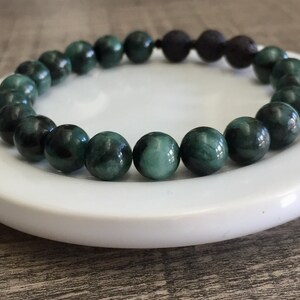 May include: A green gemstone bracelet with black lava beads. The bracelet is made of round beads and is on a white plate.