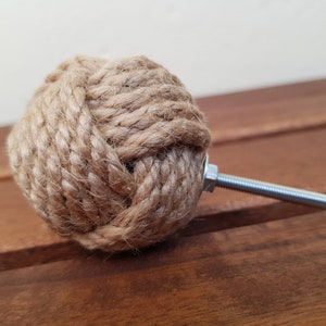 May include: A brown rope knob with a silver metal screw. The knob is tied in a knot and is a decorative accent for furniture.