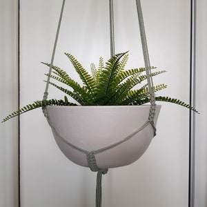 Op de afbeelding: Een witte keramische hangpot met een groene varenplant erin. De pot hangt aan een macramékoord.